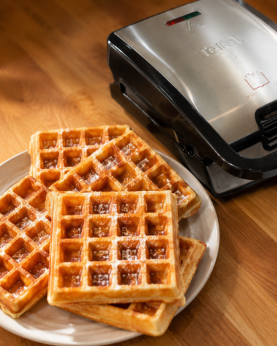 Frisch gebackene Waffeln mit Puderzucker auf einem Teller, daneben das Waffeleisen